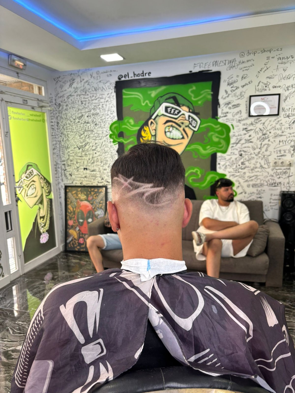 Back view of person in barbershop with geometric haircut design, facing mirror with neon green cartoon character art on wall