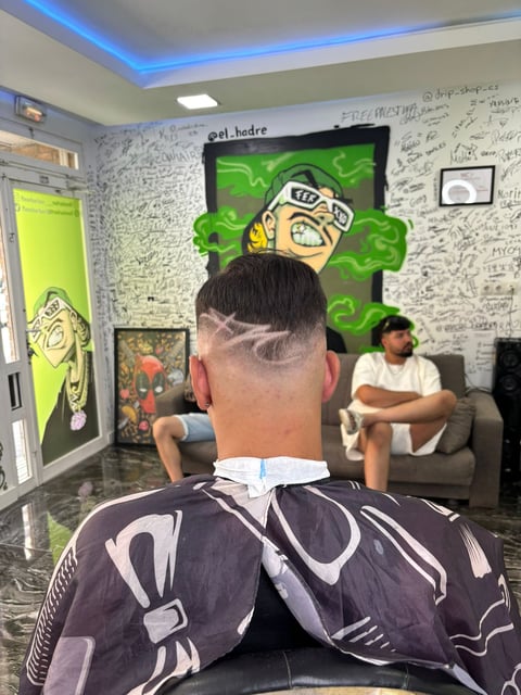Back view of person in barbershop with geometric haircut design, facing mirror with neon green cartoon character art on wall